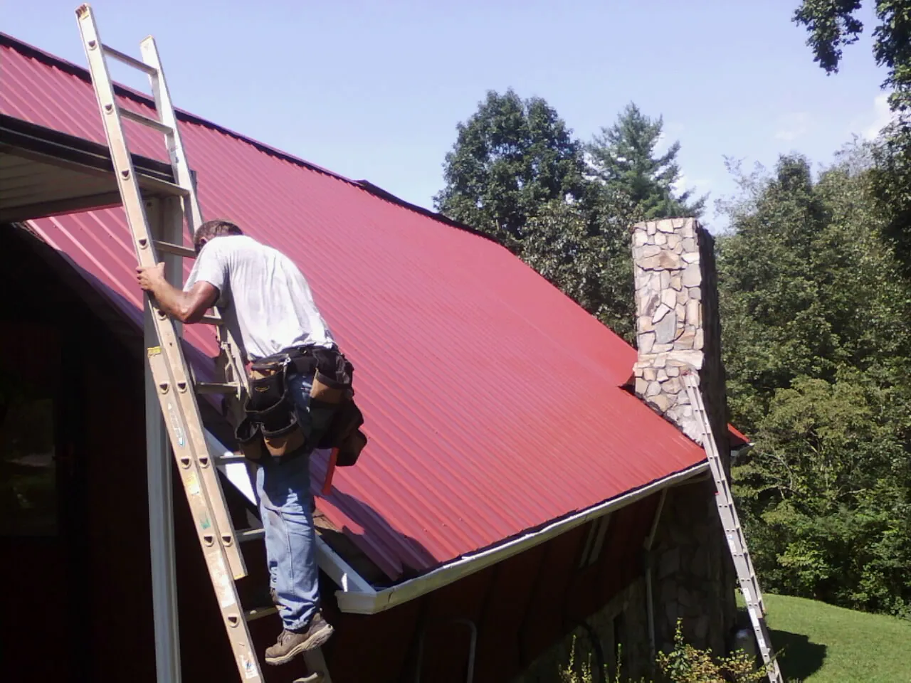 Roofer on a ladder beside a red metal roof and stone chimney — Soffit & Fascia Repair by Roofing Brothers in Tokeland